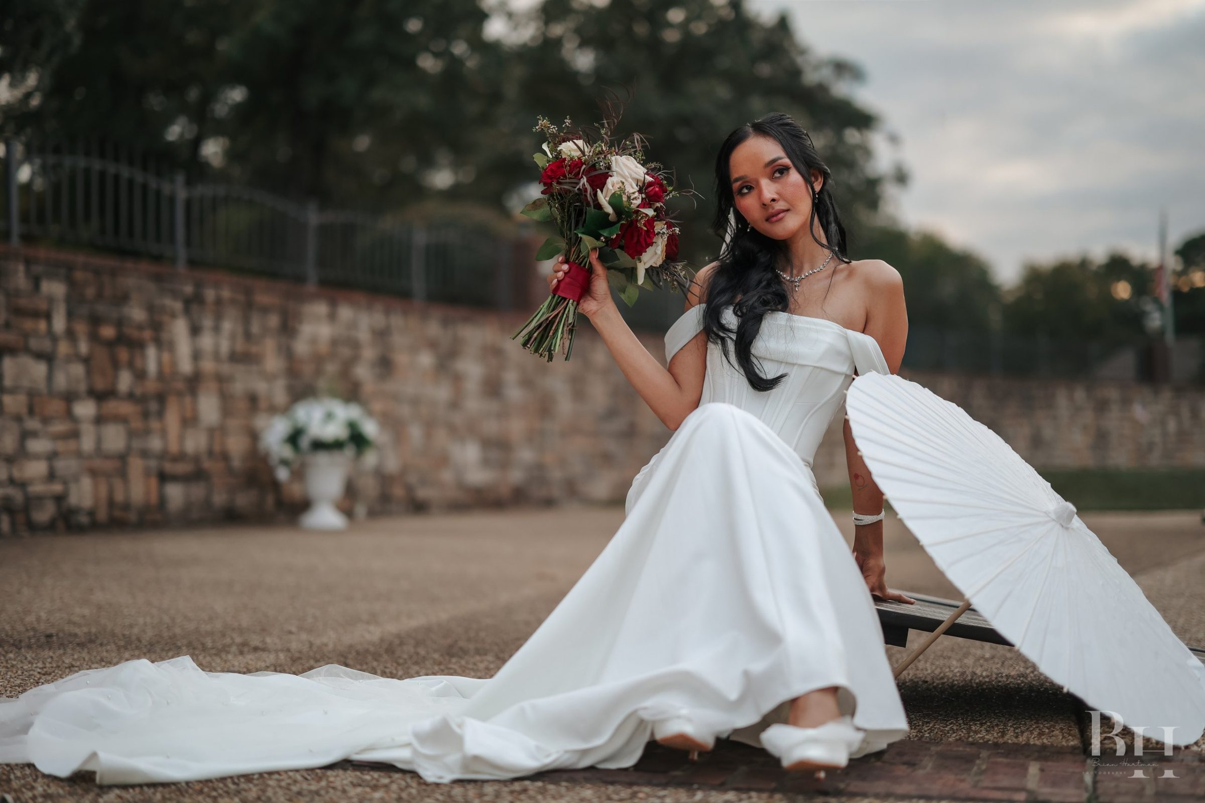 Bride in an off-the-shoulder gown seated on a stone terrace, holding a red rose bouquet beside a white paper parasol